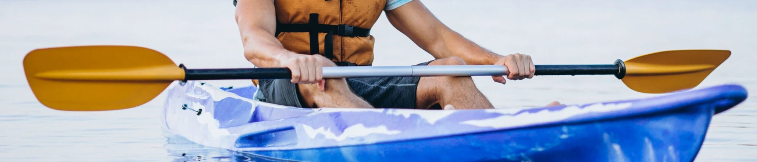A person wearing a life jacket paddling a blue and white kayak on calm water, using a double-bladed paddle with yellow blades. The lower part of their face and upper body are visible, focused on the act of kayaking.