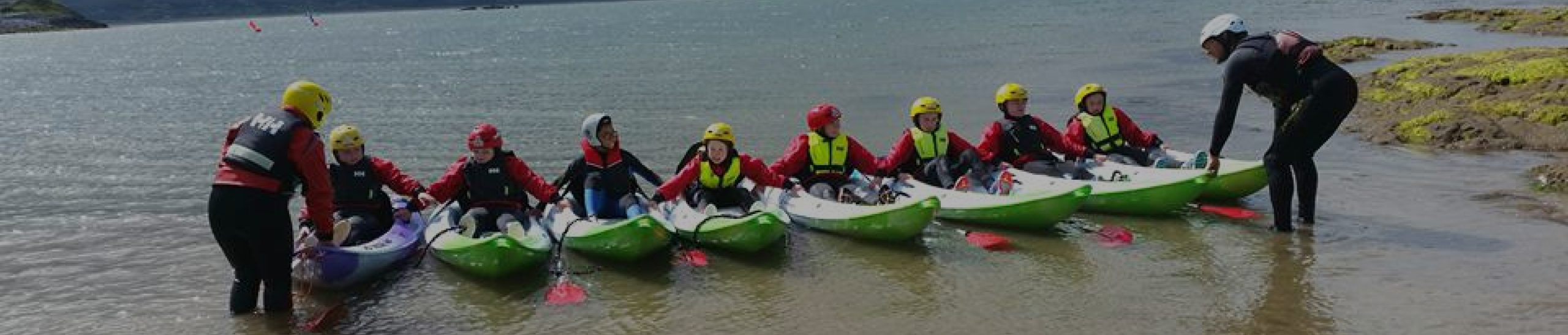 A group of nine kayakers in matching gear, including helmets and life vests, sit on their kayaks lined up at the edge of a calm body of water. An instructor leans over to assist them. The landscape includes grassy banks and distant landforms.