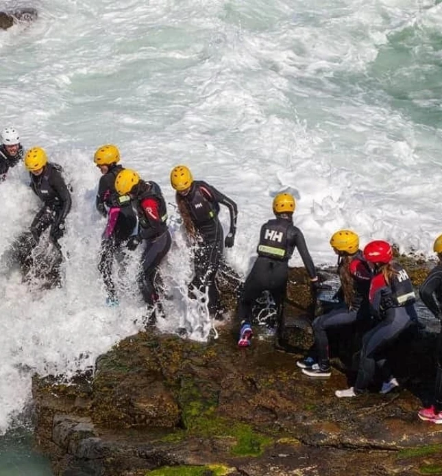 A group of people in wetsuits and yellow helmets stand on rocky terrain by the ocean, with waves splashing around them. Some are helping each other climb up the rocks while others are steadying themselves.