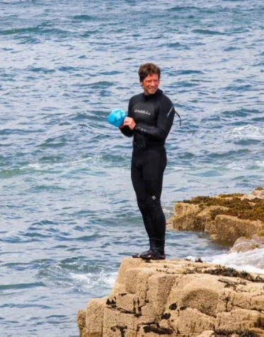 A person in a black wetsuit stands on a rocky outcrop by the ocean, holding a blue ball and smiling. Waves crash against the rocks beneath them, and the sea extends into the background.