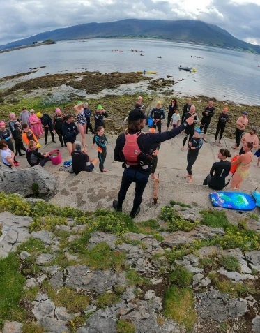 A group of people wearing wetsuits and swim gear stand on a rocky shore by a body of water, attentively listening to a person in a wetsuit holding a megaphone. In the background, small islands and hills are visible under a cloudy sky.