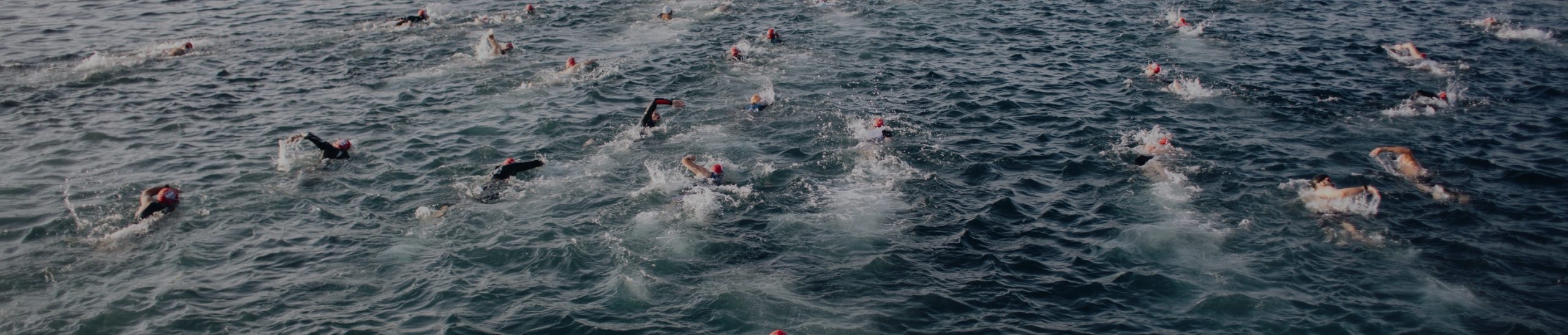 A group of swimmers with red swimming caps participating in an open water race, spread out across the choppy ocean surface. The swimmers create splashes as they move through the water. The daylight adds a blue tint to the scene.