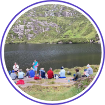 A group of people in outdoor clothing are seated on the grass near a calm lake surrounded by hills. The scene is set in a mountainous area with green vegetation and rocky terrain. One person stands while the others are seated, possibly listening or taking a break.