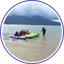 A group of six people in kayaks is being instructed by a person standing in the shallow water by the shore. The kayaks are aligned side by side on a calm sea with mountainous terrain and cloudy skies in the background. Everyone is wearing life jackets and helmets.