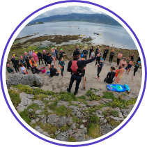 A group of people in wetsuits stand on a rocky beach near the water, listening to a person giving instructions. The shoreline is uneven, with patches of grass and rocks. The background shows a cloudy sky and distant hills.