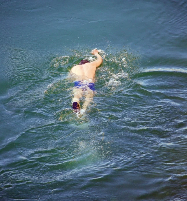 A person wearing blue swim shorts is swimming in a large body of water, performing a freestyle stroke with one arm outstretched and the other close to their body, creating ripples and splashes in the water.