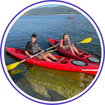 Two people are sitting in red kayaks on a calm lake. They are holding yellow paddles and wearing life jackets. The lake is surrounded by distant mountains, and the sky is clear. The scene is peaceful and bright.