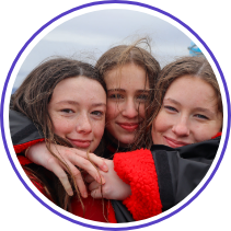 Three young women with windblown hair huddle close together outdoors, wearing thick red and black jackets. They smile contentedly, with their faces touching, suggesting a close bond or friendship. The background is an overcast sky, indicating a cool or breezy day.