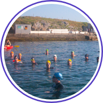 A group of people wearing colorful swim caps are floating and swimming in open water near the shore. The background features a rocky coastline with buildings and structures, and the event appears to be a group swimming activity.