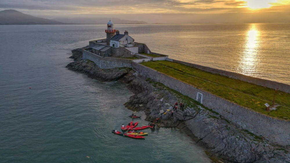 A group of people with colorful kayaks gather on rocky shore near a stone lighthouse on a coastal peninsula at sunset, with calm water and distant mountains in the background.