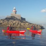 Two people in red kayaks paddle near a rocky island with a lighthouse on top. The water is calm and the sky is clear with a few clouds.