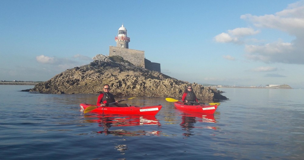 Two people in red kayaks paddle near a rocky island with a lighthouse on top. The water is calm and the sky is clear with a few clouds.