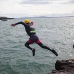 A person wearing a helmet and wetsuit jumps off a rocky edge into the sea, while another person in similar gear stands nearby and a third person waits in the water.