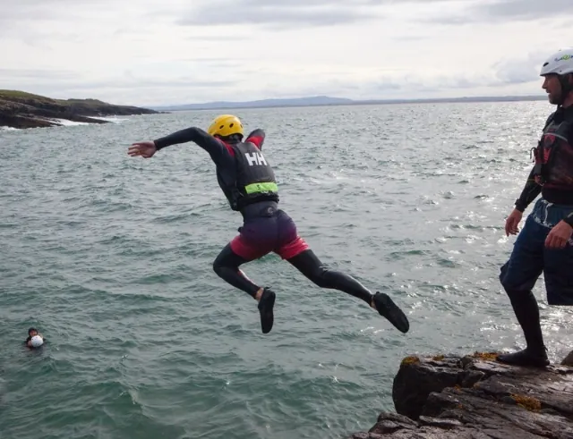 A person wearing a helmet and wetsuit jumps off a rocky edge into the sea, while another person in similar gear stands nearby and a third person waits in the water.
