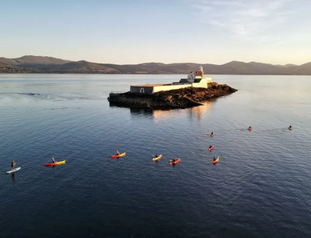 A small rocky island with a lighthouse sits in calm water, surrounded by mountains. Several people in kayaks and on paddleboards are paddling nearby under a clear sky.