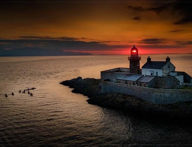 A lighthouse with a bright red light stands on a rocky coastline at sunset, overlooking calm sea waters. Kayakers are visible paddling in the distance under the orange and red sky.