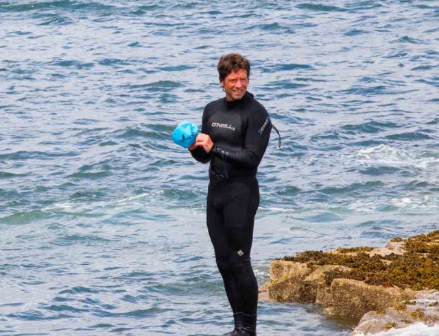 A person in a black wetsuit stands on a rocky shore, holding a blue swimming cap, with waves from the sea in the background.