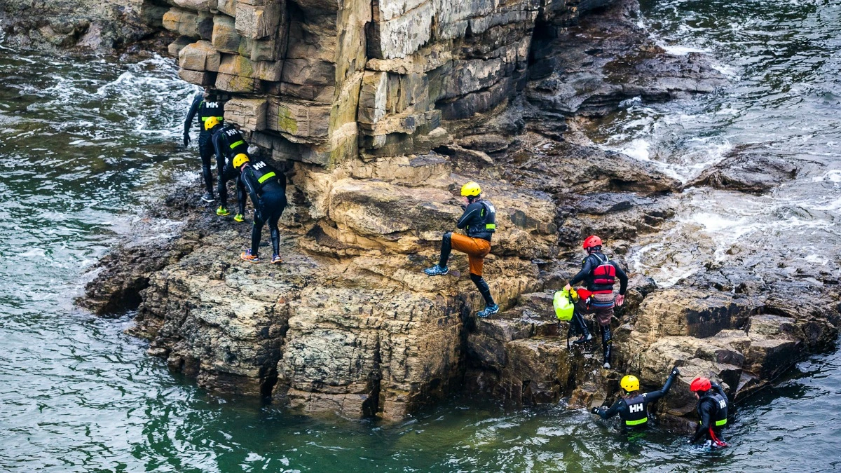 climbing rocks around sea A group of people in wetsuits and helmets climb and walk on rocky cliffs beside the sea, with waves splashing at their feet, engaging in a coastal outdoor adventure.